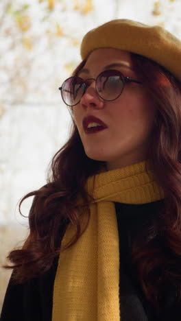 dama elegante con gafas de sol, boina y silenciador amarillo mira los pilares de madera decorativos, rodeados de follaje de otoño con hojas doradas en el fondo, evocando una atmósfera elegante y tranquila