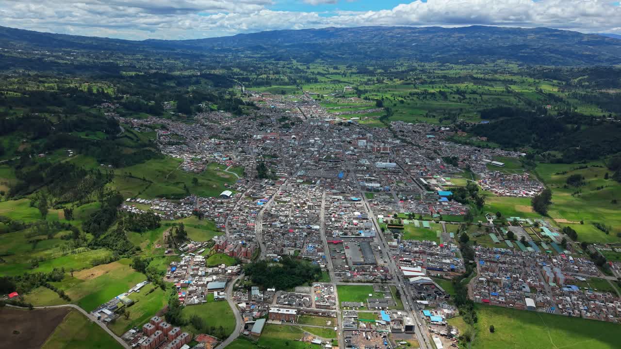 High drone pullback revealing the full city of Chiquinquirá, with moving cloud shadows, surrounding green fields, and distant Colombian mountains