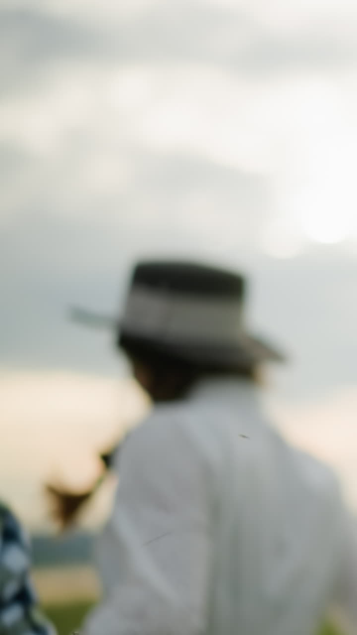 задний вид пары, держащейся за руки и бегущей по высокому травяному полю. мужчина в черной шляпе и клетчатой рубашке, в то время как женщина носит белое платье и шляпу, захватывая радостный