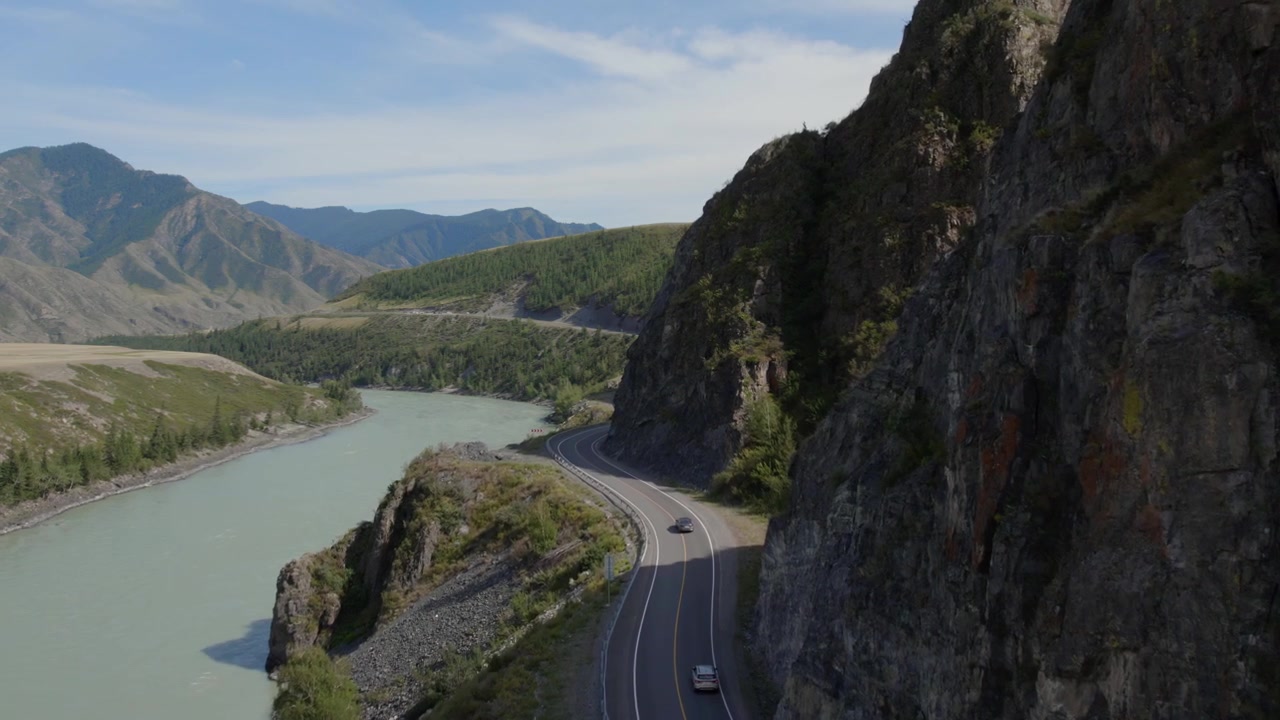 Vista aerea di auto che percorrono la tortuosa Chuysky Trakt accanto al fiume Katun turchese, circondate da scogliere rocciose e montagne nella Repubblica dell'Altai, Russia.
