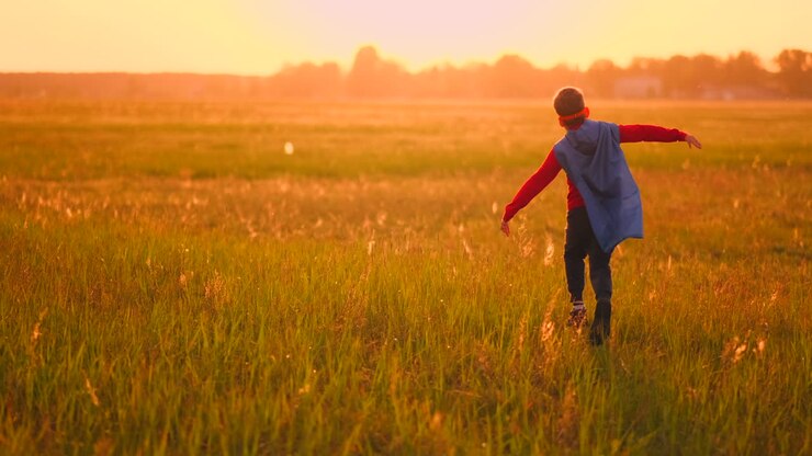 A boy in a suit and a superhero mask with a red cloak runs across the field at sunset on the grass dreaming and imagining himself a hero and a winner.