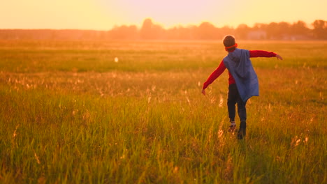 Ein-Junge-In-Einem-Anzug-Und-Einer-Superheldenmaske-Mit-Rotem-Umhang-Läuft-Bei-Sonnenuntergang-über-Das-Feld-Auf-Dem-Gras-Und-Träumt-Und-Stellt-Sich-Vor,-Er-Sei-Ein-Held-Und-Gewinner.