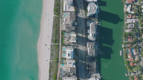 Top-down-view-of-traffic-on-wide-road-lined-with-tall-buildings-in-urban-neighbourhood.-Narrow-stripe-of-land-surrounded-by-turquoise-water.-Miami,-USA