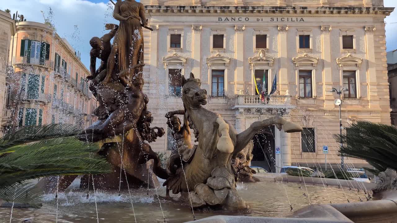 Early afternoon view of the Fountain of Diana in Ortigia, Sicily, with water flowing and the Banco di Sicilia building behind, showcasing classical sculpture and historic urban architecture