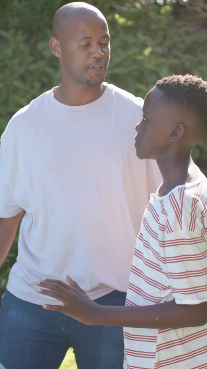 Video verticale: Papà in maglietta che calma un adolescente reagendo a un gesto, aprendo le braccia in giardino, con impugnatura arancione.