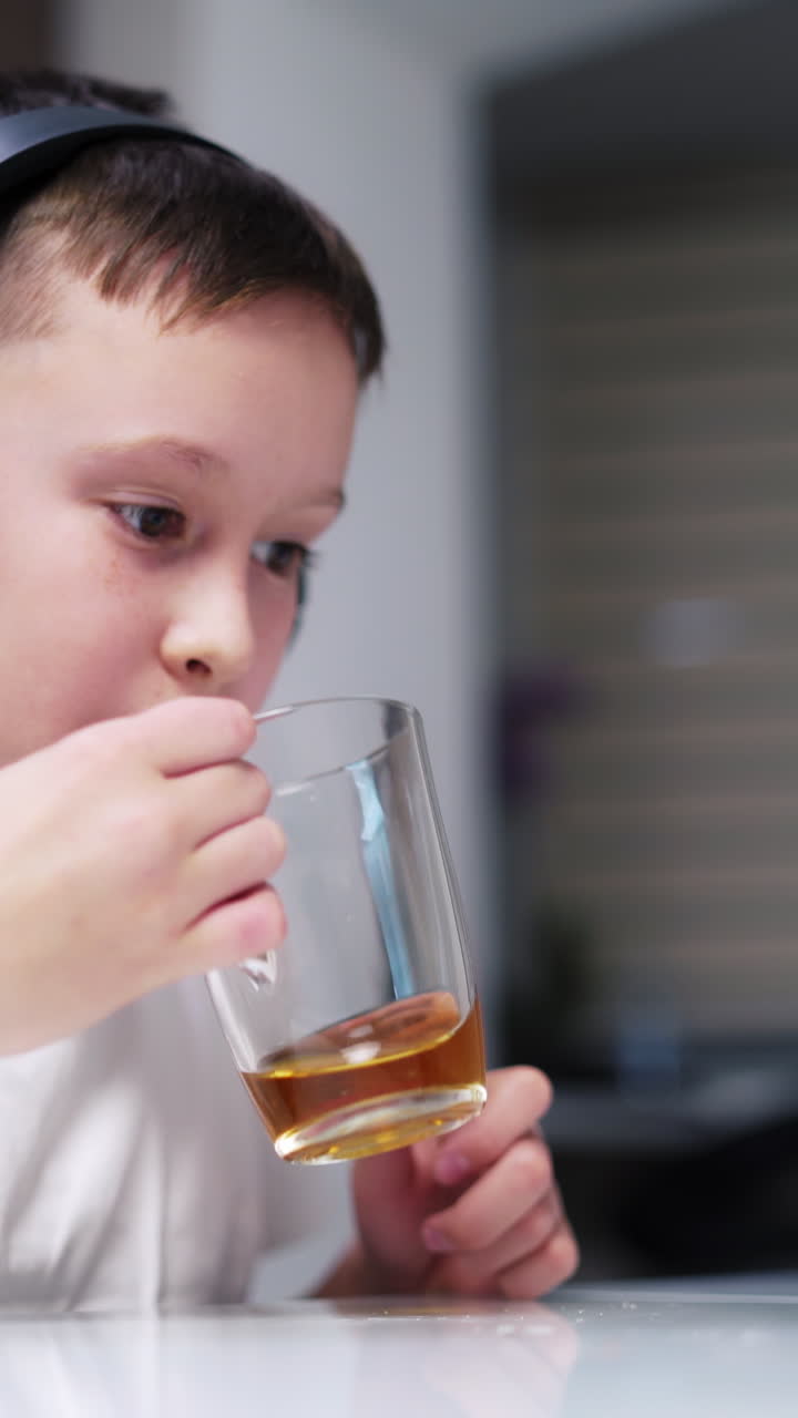 Boy in headphones playing on tablet and drink. Concentrated on game teenage boy is sitting at the table and drinking tea at home. Vertical video