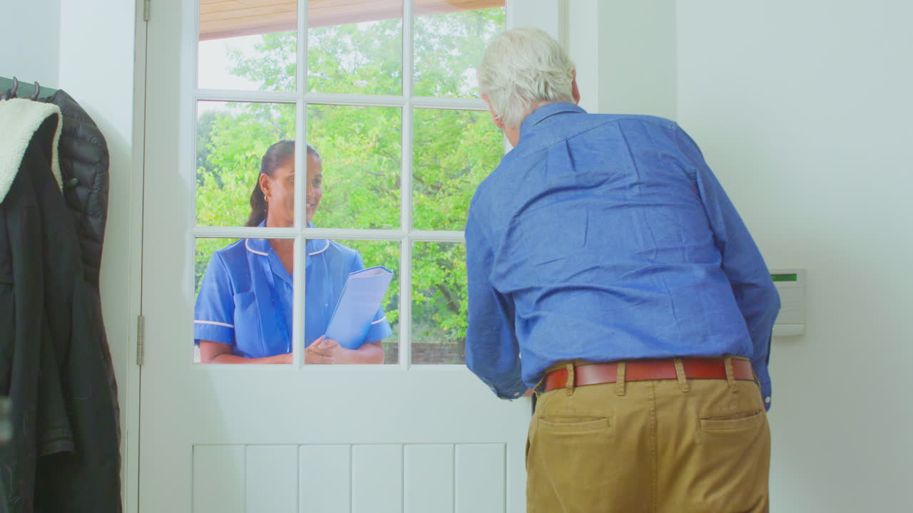 Senior Man At Home Using Walking Stick Greeting Female Nurse Or Care Worker In Uniform At Door