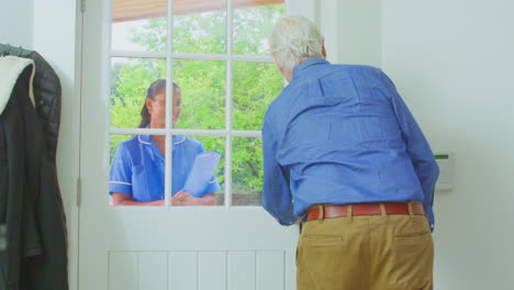 senior man at home using walking stick greeting female nurse or care worker in uniform at door