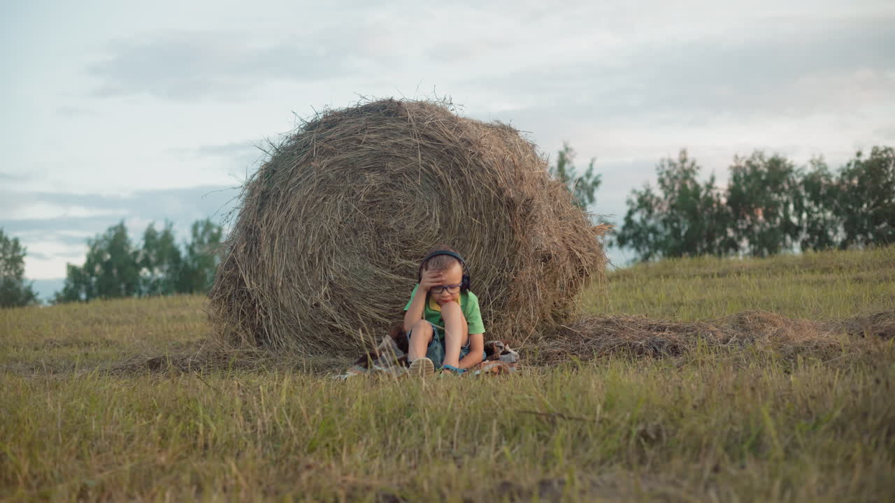 маленький мальчик в зеленой рубашке, наушниках и очках, спирается головой на колени, рукой на голову, сосредотачиваясь на музыке, на фоне обширных сельскохозяйственных угодий