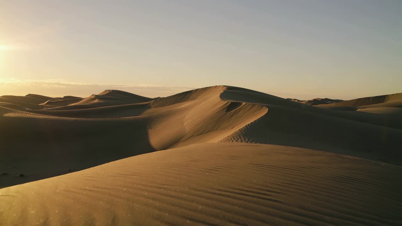Telecamera in movimento all'alba sopra una duna a mezzaluna che evidenzia la cresta mentre il sole mostra increspature e granelli.