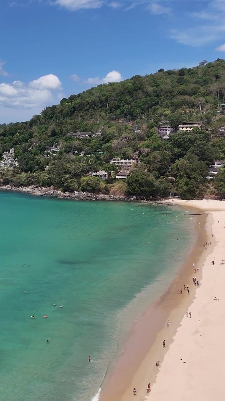 Un filmato di drone cattura una spiaggia serena a Phuket, Thailandia, mostrando acque turchesi, spiagge sabbiose e una vegetazione lussureggiante sotto cieli sereni.