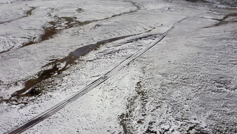 Antena:-Siguiendo-Un-Coche-Blanco-En-Un-Camino-De-Tierra-Embarrado,-Parcialmente-Cubierto-De-Nieve-En-Las-Tierras-Altas-De-Islandia