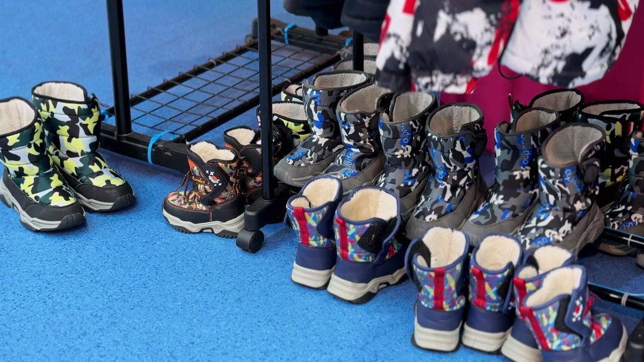 Eine Reihe warmer Kinder-Winter-Schneestiefel mit Tarnmuster, aufgereiht auf einem blauen Boden in Innenräumen. Das Schuhwerk steht unter einem Kleiderständer in einem Verleihgeschäft für Wintervergnügen unter heller Beleuchtung.