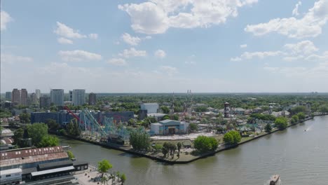 aerial drone view: amusement park with roller coasters beside tranquil river in tigre, argentina. lush greenery surrounds waterfront attractions under cloudy sky, showcasing leisure destination