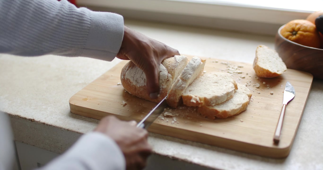 African American man holding loaf, slicing bread with bread knife on board by window preparing food