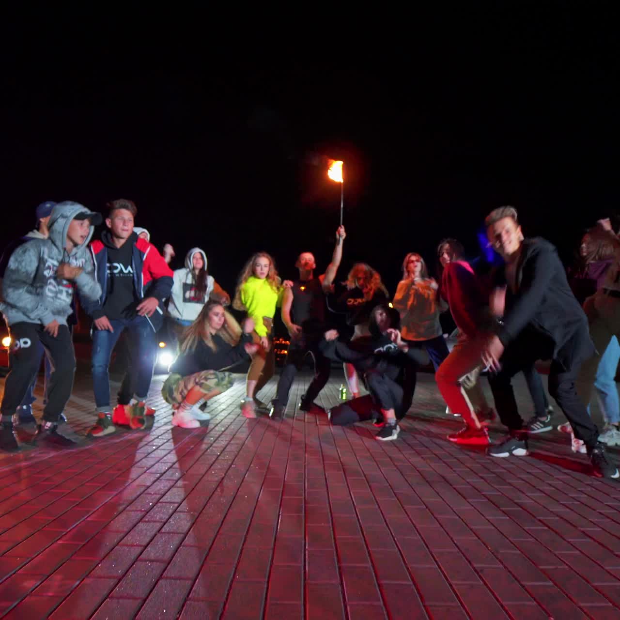 Group of teenagers standing in half-circle dance hip-hop. Young man in the middle holding a lit torch. Night square backdrop