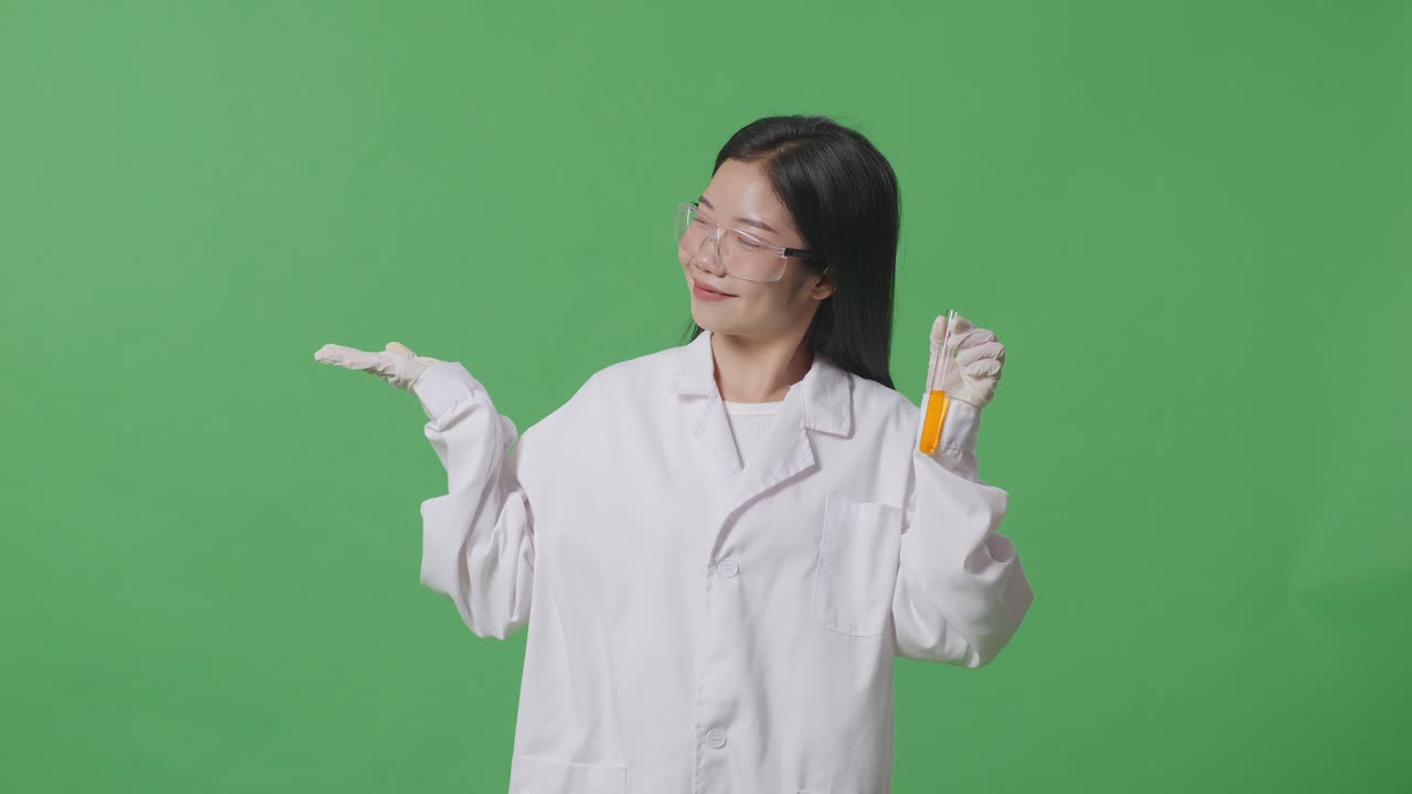 실험실에서 녹색 화면 배경에 서 있는 동안 미소 짓고 으로 가리키는 시험관에서 오렌지 액체와 함께 아시아 여성 과학자
