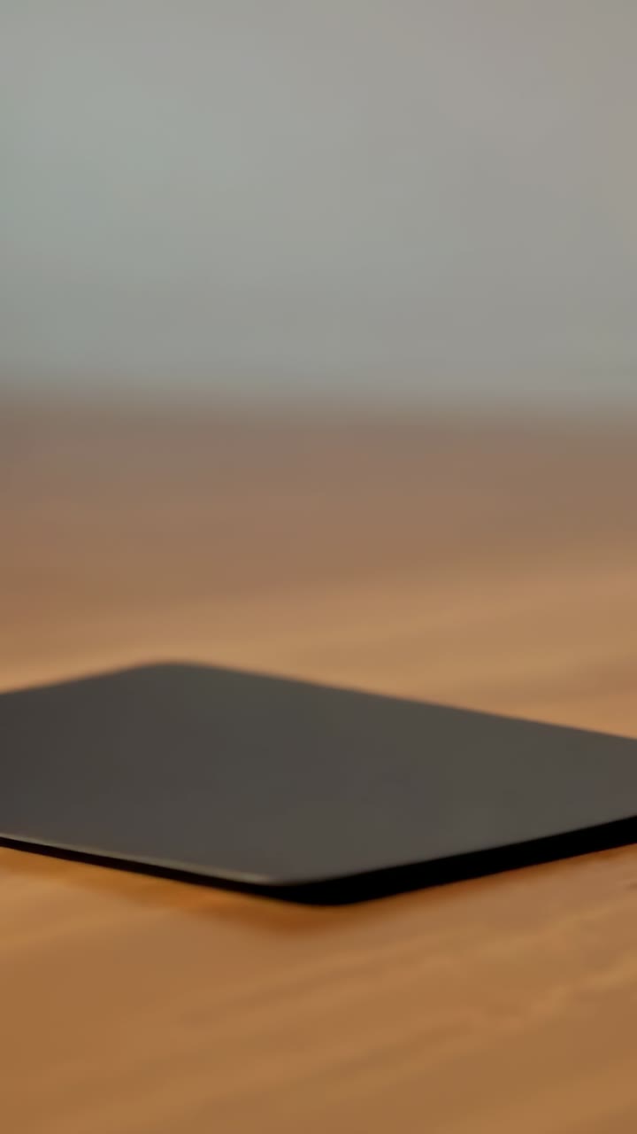 Vertical video: Sliding thin dark pad tilting, settling onto wood table while hidden arm placing