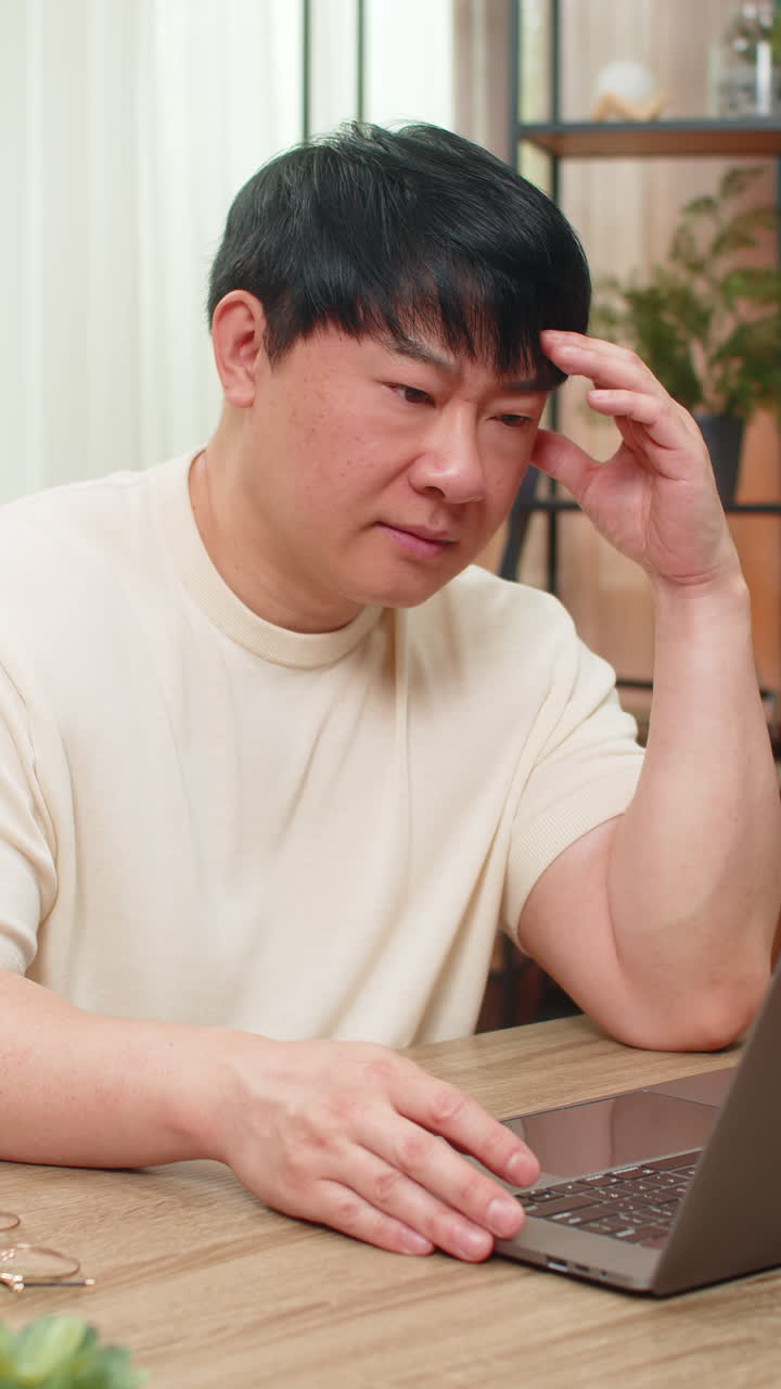 Asian man angrily rereading email on laptop tapping keyboard tense face after receiving bad news