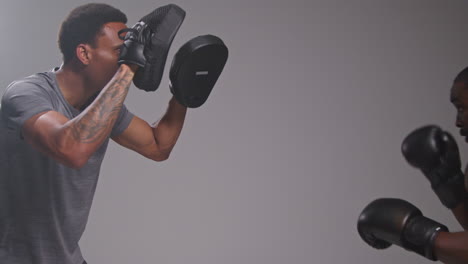 Studio-Shot-Of-Male-Boxer-Sparring-Working-Out-With-Trainer-Wearing-Punch-Mitts-Or-Gloves-Practising-For-Fight-1