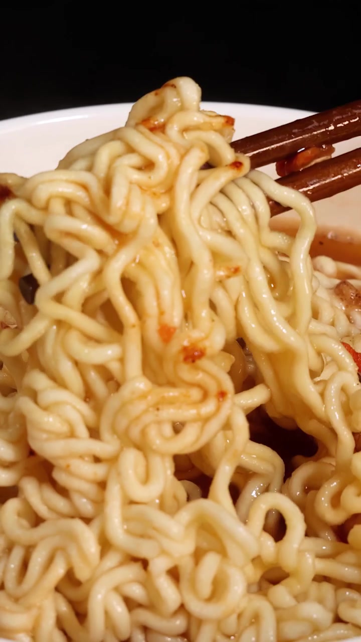 Le bacchette afferrano e sollevano noodles istantanei cotti da una ciotola fumante di brodo. Primi piani, illuminazione calda e uno sfondo scuro mettono in risalto la consistenza e il movimento