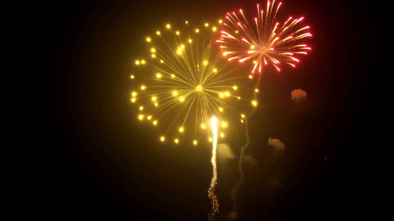 Colorful fireworks erupt high in dark sky with bright red and golden bursts, radiant spark trails, glowing embers, dense smoke, and vivid light creating energetic festive atmosphere