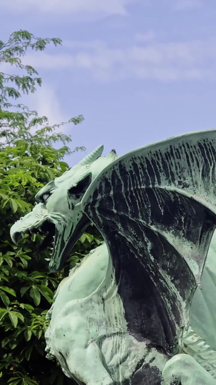 Primo piano verticale dell'iconica scultura del drago in rame verde sul Ponte dei Draghi a Lubiana, Slovenia, incorniciata da alberi frondosi e cielo blu