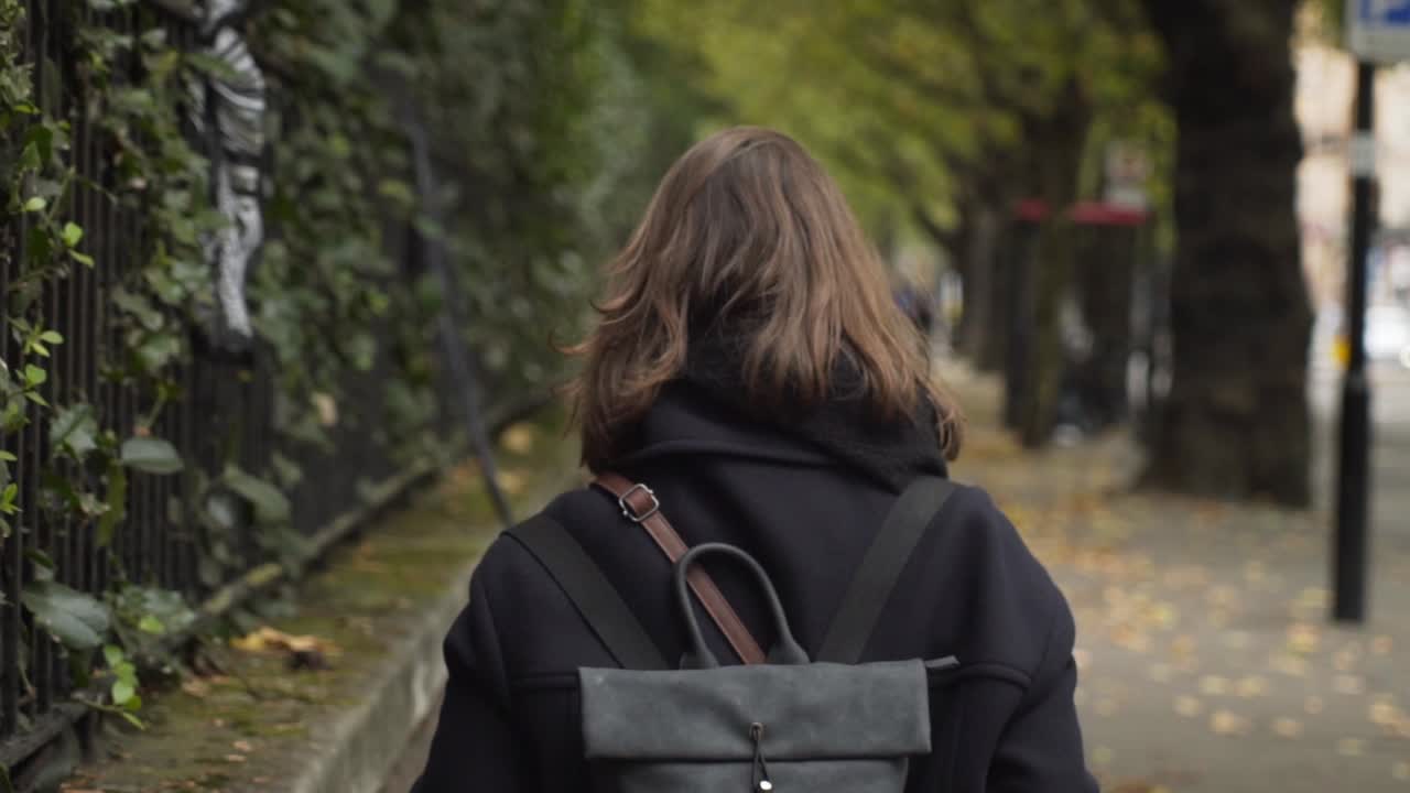 녹색 초목과 잎이 있는 강철 연철 울타리를 따라 런던 시멘트 보도를 걷고 있는 젊은 백인 여성의 아름다운 가을 전망, 클로즈업 슬로우 모션 뒤에 핸드헬드