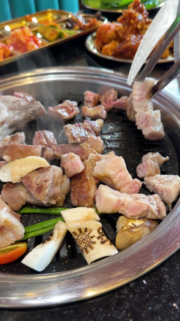 Une main utilise des pinces pour couper de la poitrine de porc sur un barbecue coréen grésillant, sous un éclairage intérieur lumineux.