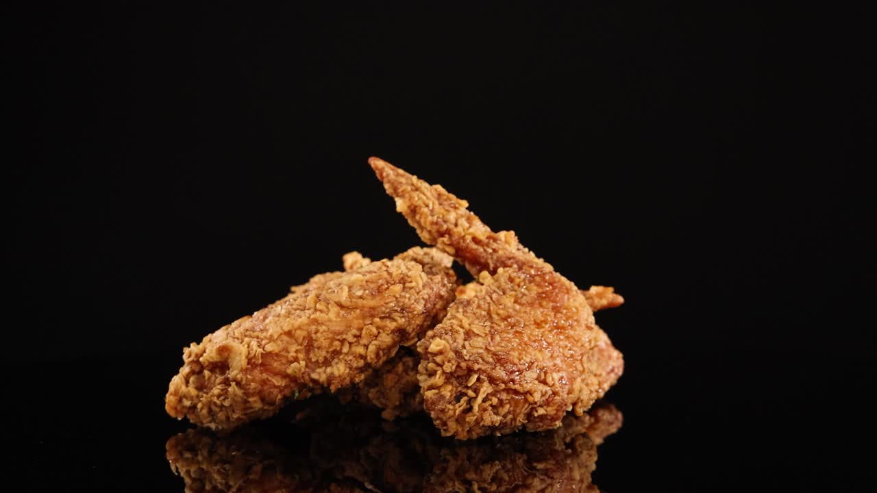 Golden-brown fried chicken wings rotate smoothly on a reflective black surface, illuminated by studio lighting for a dramatic, appetizing presentation with seamless camera movement