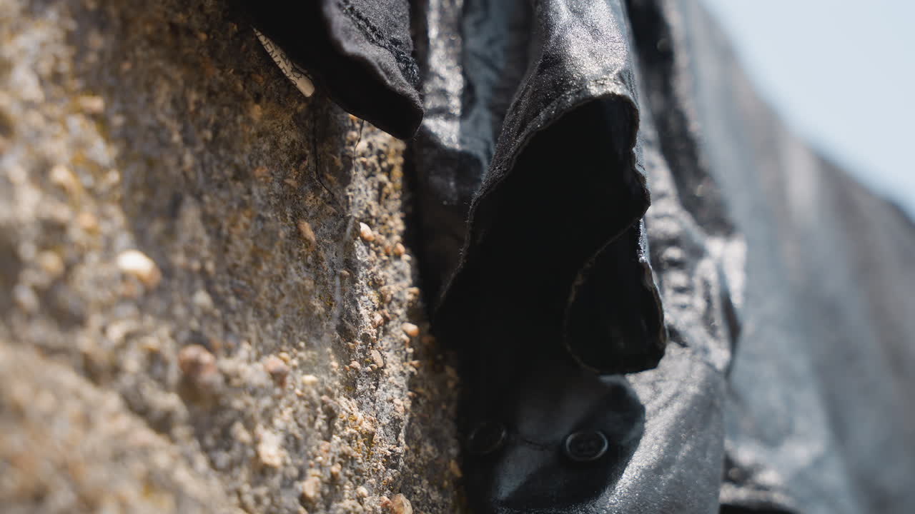 Black garment drying on stone wall crevice, closeup of damp sleeve tucked into rough mortar, gritty pebble detail and weathered texture, domestic laundry chore captured in muted light