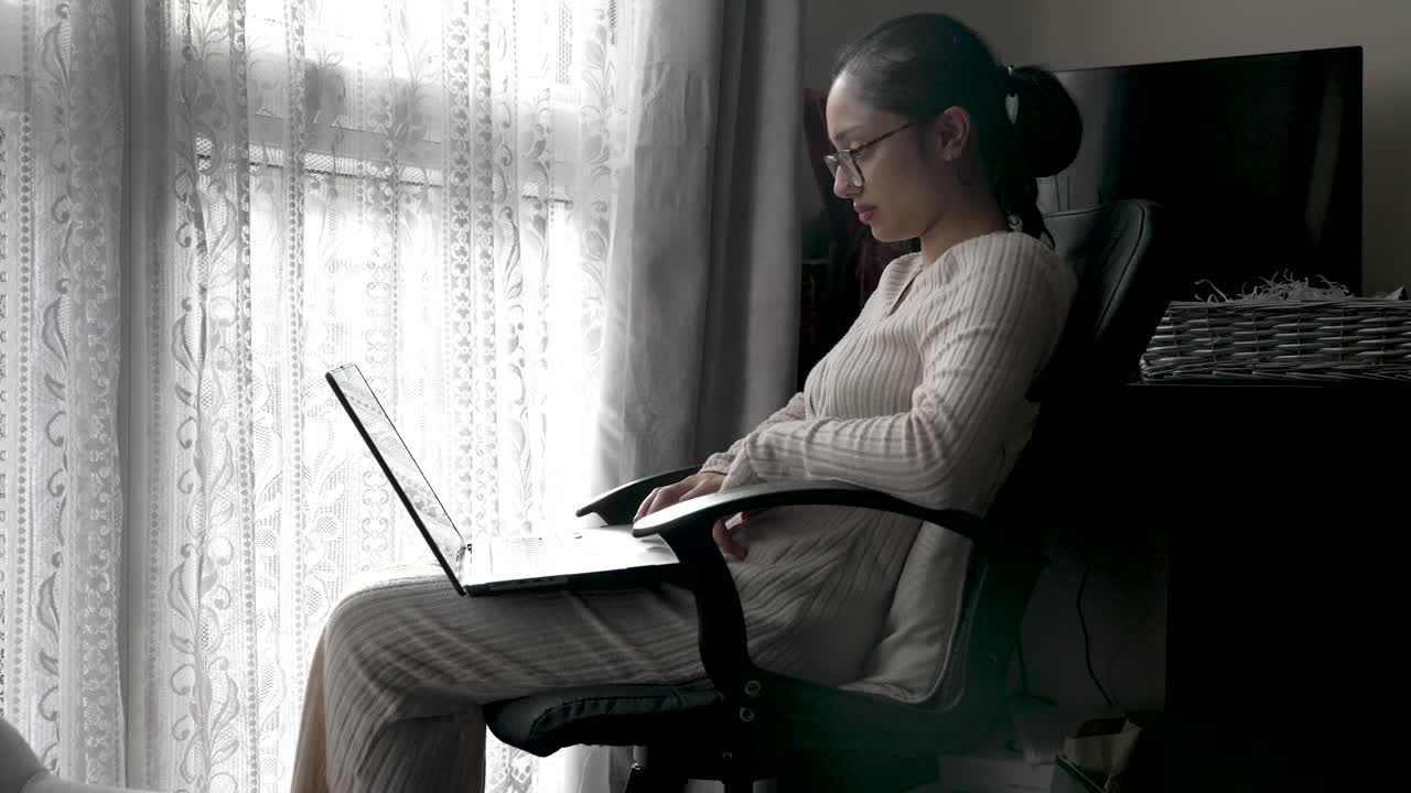 una persona se sienta cerca de una ventana, trabajando en una computadora portátil colocada en su regazo, mostrando la integración de los medios digitales en entornos domésticos, para entretenimiento multimedia o actividades educativas