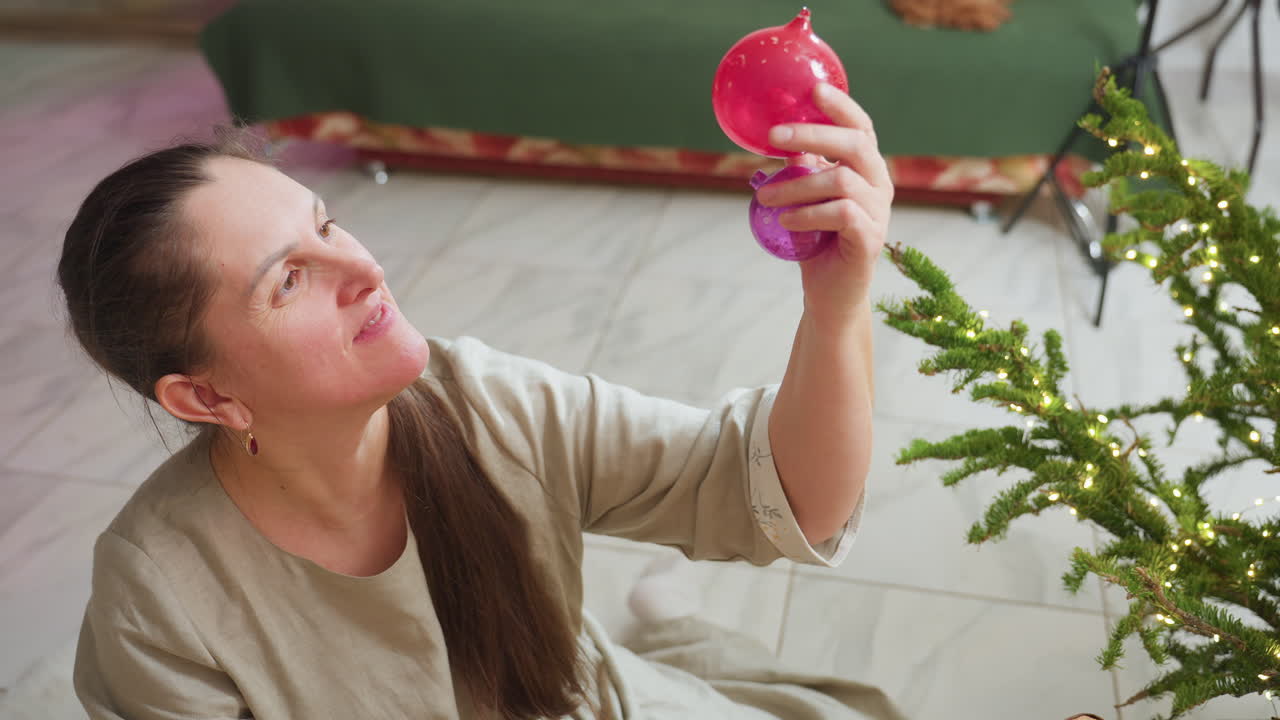 Woman sitting on floor holding two colorful Christmas ornaments in hand, inspecting them closely with joyful expression near decorated Christmas tree during cozy festive holiday season indoors