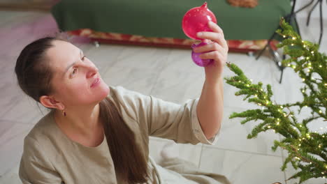 woman sitting on floor holding two colorful christmas ornaments in hand, inspecting them closely with joyful expression near decorated christmas tree during cozy festive holiday season indoors