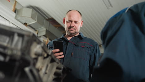 lower angle view of mechanic in blue uniform working on engine with red hand glove, while colleague nearby operates phone in automotive workshop