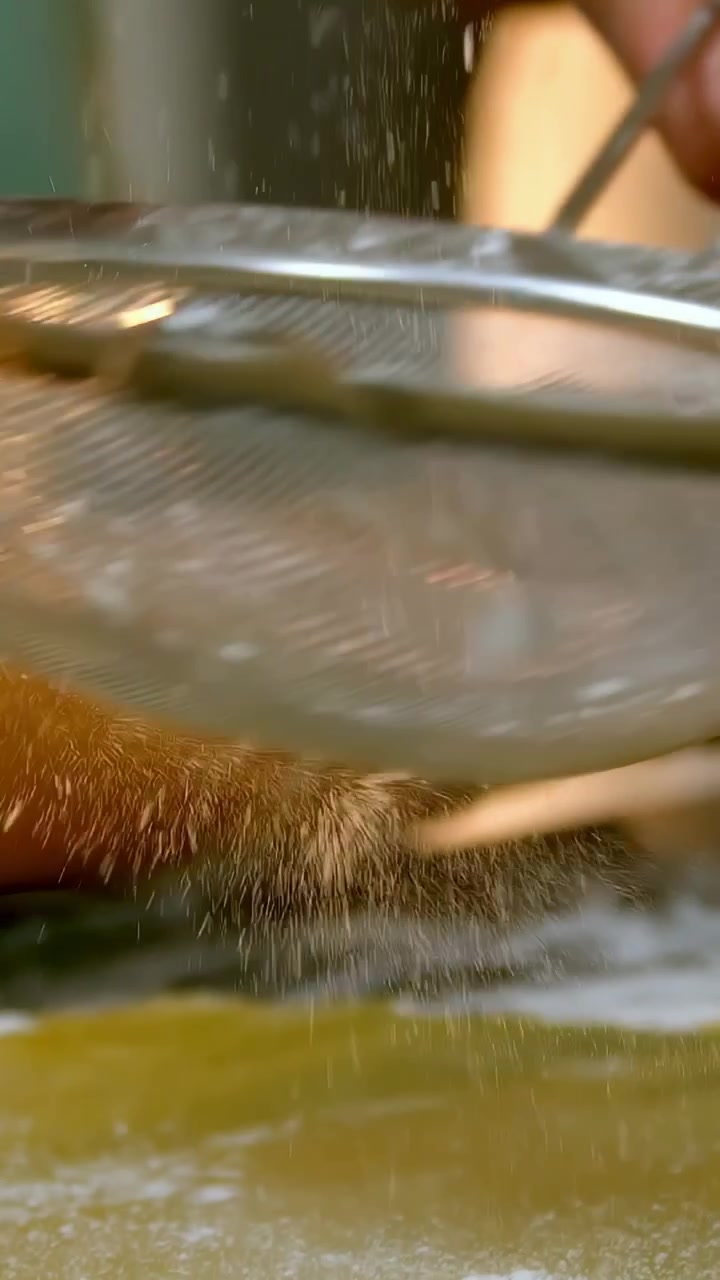 Männliche Hand im grünen Oberteil bestäubt Teig gleichmäßig mit einem Sieb auf der Küchenarbeitsplatte, daneben ein Nudelholz.