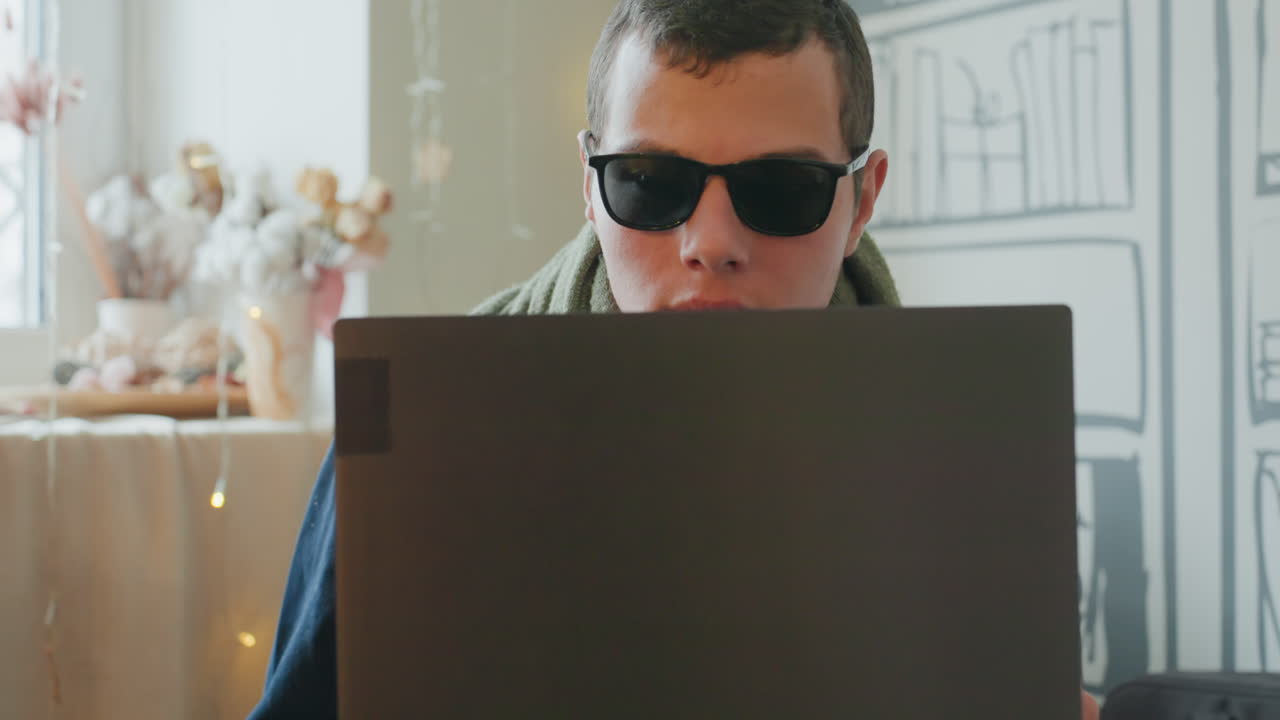 Uomo bianco con occhiali da sole dietro il laptop, ambiente accogliente del soggiorno, decorazioni floreali, lucine fatate, postura casual da freelancer, mani sulla tastiera, espressione guardinga, atmosfera di lavoro a distanza.