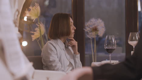 serious woman sitting alone at restaurant table and drinking wine while female musician playing piano