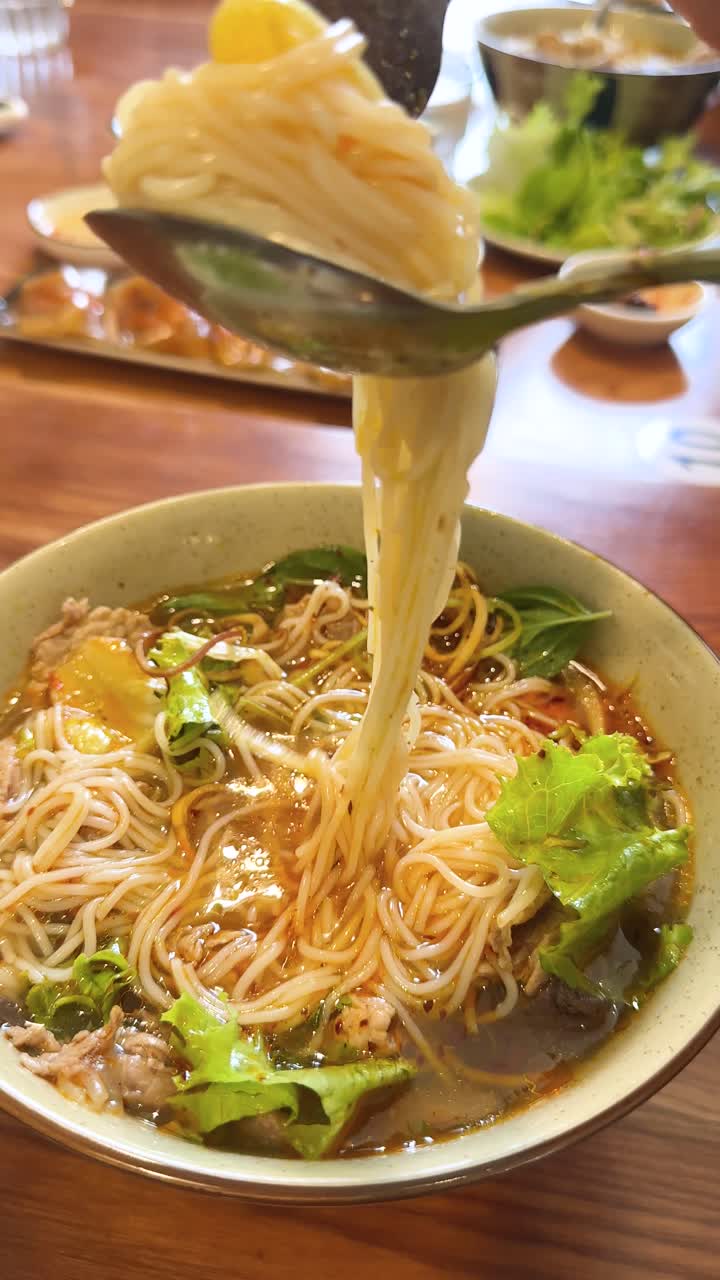 Primo piano di mani che usano una forchetta e un cucchiaio per sollevare noodles da un brodo piccante.