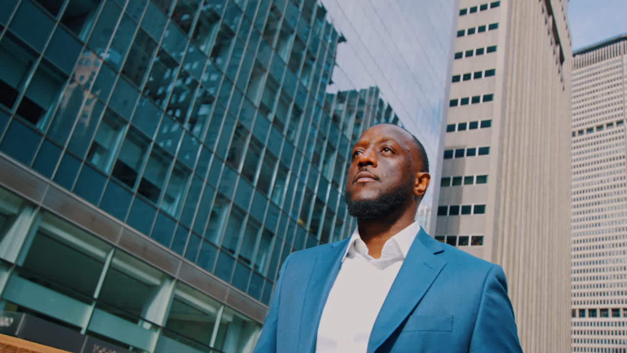 New York, USA, 8 April 2026: American businessman standing against glass skyscraper in Manhattan. Low angle shot of Black man in blue suit looking away with modern glass office building background