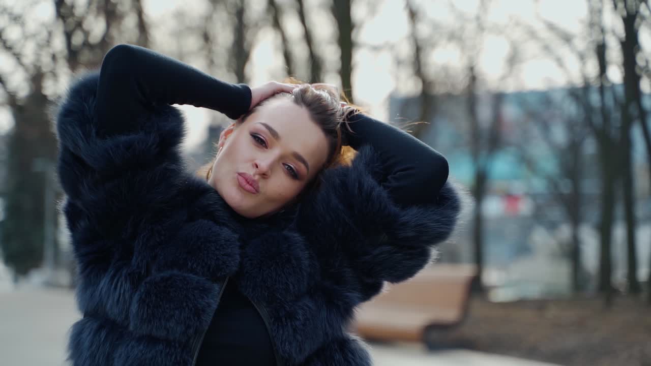 Portrait of a woman outdoors. Sexy girl in black fur coat looking on camera while holding her brown hair on the city park background. Slow motion.