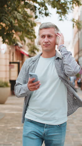 young grayhaired man listening to music using wireless headphones and smartphone on city street