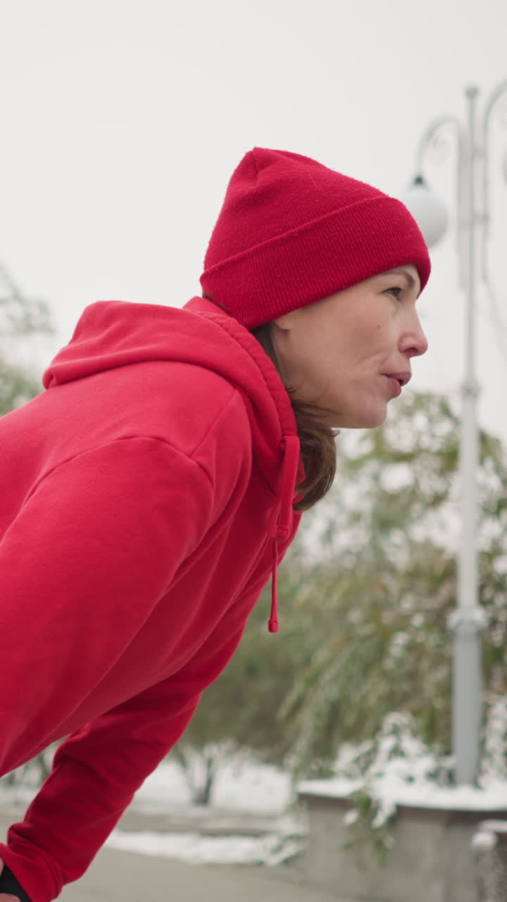 Woman in red hoodie and beanie bends down resting, gazing into distance, snowy background with lamp post, frosted trees, benches, iron railing, and mast in foggy winter atmosphere