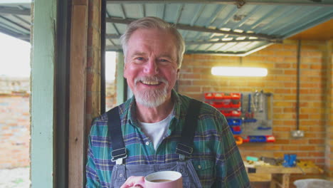 Portrait-Of-Senior-Male-Wearing-Overalls-In-Garage-Workshop-With-Hot-Drink