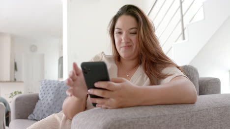 mujer biracial feliz de tamaño plus teniendo llamada de video de teléfono inteligente sentada en el sofá en casa, cámara lenta