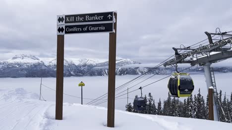 Señal-De-Pista-De-Esquí-De-Diamante-Negro-Doble-En-La-Parte-Superior-De-La-Estación-De-Esquí-De-Revelstoke-Mt-Mackenzie-Con-Gondalas-En-Movimiento-En-El-Fondo-Y-Una-Vista-De-Las-Montañas-Nevadas