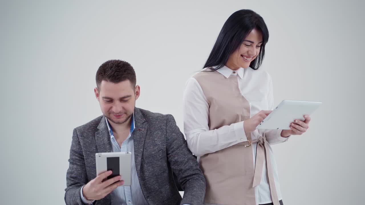 Young couple with modern devices in studio. Smiling woman looking at digital tablet. Handsome man sitting in chair and watching video on tablet.