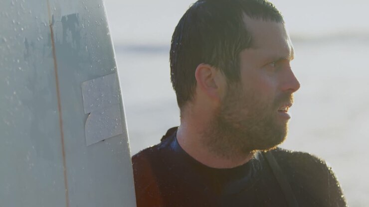 Side view of mid-adult caucasian male surfer with surfboard standing at beach during sunset 4k