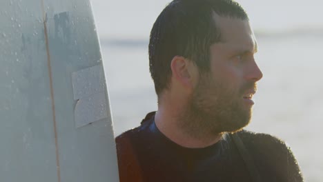 Side view of mid-adult caucasian male surfer with surfboard standing at beach during sunset 4k