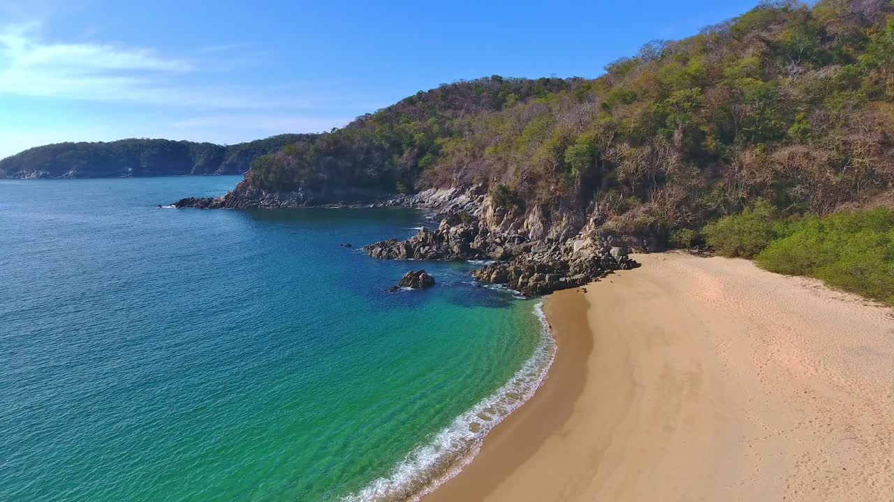 Volo stazionario in avanti a bassa angolazione di una vista sulla spiaggia con piccole onde che si infrangono vicino a una costa rocciosa e cristallina
